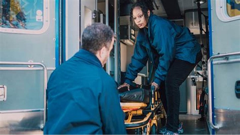 Two people in blue uniforms loading an empty stretcher into an ambulance, with medical equipment visible inside.